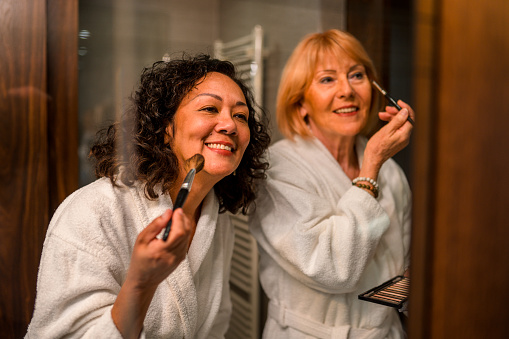 Two older women wearing white robes are doing their make-up together in their hotel room. A smiling Hispanic woman with curly brown hair is holding a make-up brush towards her face. A Caucasian woman with short blonde hair is smiling while applying eye shadow. Friends getting ready for girl's night out.