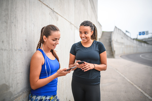 Two sporty and beautiful young women laughing and socialising while holding their smart phones. They are wearing fitness clothing and leaning on a concrete wall.