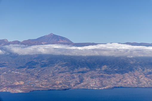 View to the coast of Tenerife with a wind turbine park and the peak of the volcano Teide above a layer of clouds seen from an airplane