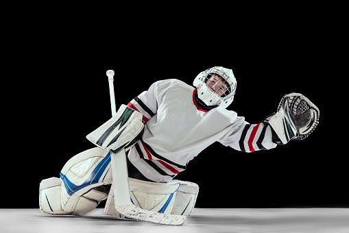Portrait of teen boy, hockey player, goalkeeper catching plug in motion isolated over black studio background. Dynamic sport. Concept of sportive lifestyle, childhood, hobby, action