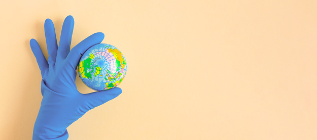 Earth globe in hands in blue protective glove on a clean beige background. The concept of protecting nature, ecology and global peace. World globe in Hand. The COVID-19 pandemic. Theme of earth day. Green planet earth, held by hands in gloves. Caring for the climate, nature, ecology. Flat lay for websites, magazines, apps. Hands holding globe on a beige background.