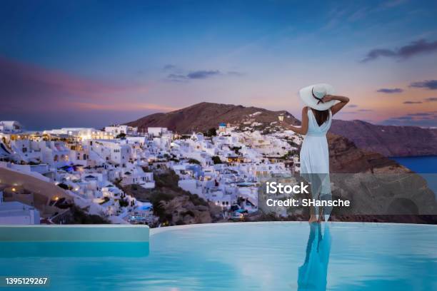 A Stands By The Swimming Pool And Enjoys The Sunset View Over The Illuminated Village Of Oia Stock Photo - Download Image Now