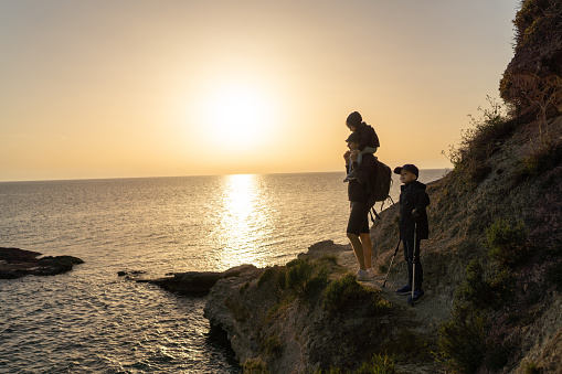 Father standing with son and little daughter on his shoulders and enjoying sunset while taking a break from hiking on the rocks by the sea