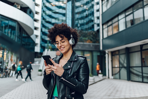 Young stylish pretty woman with afro hair walking in the city streets and using smartphone and headphones. Female hipster reading message on mobile and listening songs from a playlist.