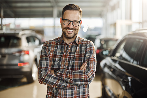 Happy salesperson standing in a car showroom with his arms crossed and looking at camera.