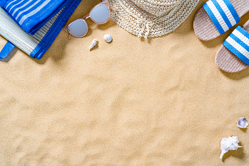 Summer backgrounds: beach sandals, sunglasses, beach towel, beach mat and sunhat shot from above on sand background. The composition is at the top of an horizontal frame leaving useful copy space for text and/or logo. High resolution 42Mp outdoors digital capture taken with SONY A7rII and Zeiss Batis 40mm F2.0 CF lens accessories