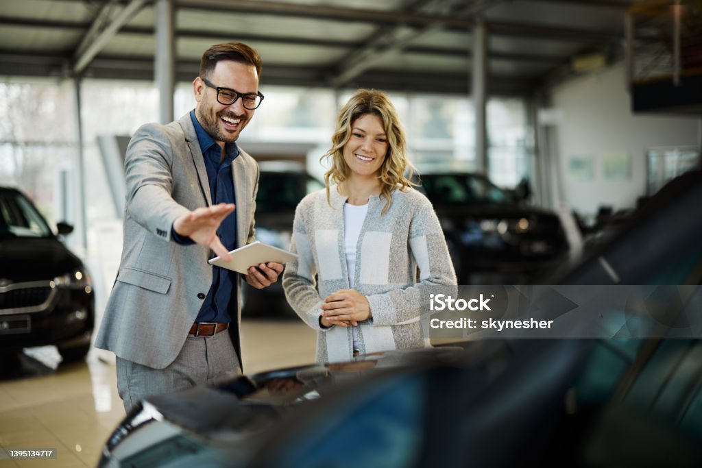 Happy salesman selling the car to his female customer in a showroom. Happy car salesman showing a car to his customer in a showroom. Car Salesperson Stock Photo