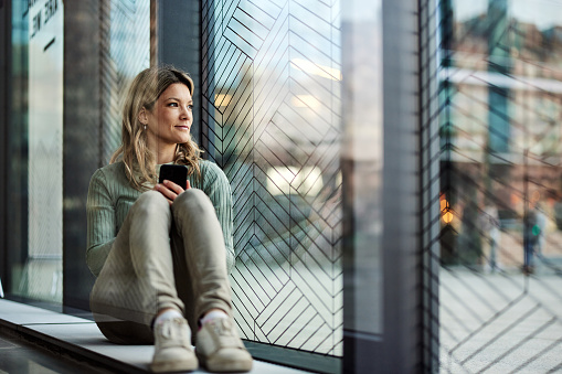 Young smiling woman day dreaming while using smart phone by the window.