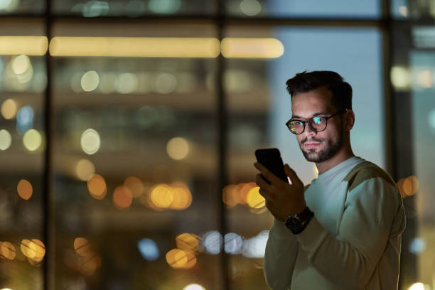 Young serious man reading a text message on smart phone in the dark. Young man text messaging on cell phone indoors at night. guy looking at cell phone stock pictures, royalty-free photos & images