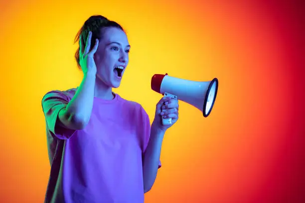 Attention. Beautiful excited girl shouting at megaphone isolated on yellow-red color background in neon light, filter. Concept of emotions, expression, youth, aspiration, sales. Copy space for ad