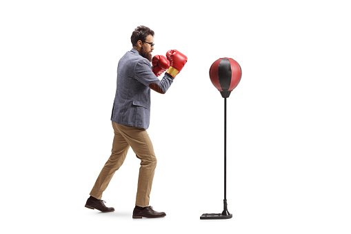 Full length profile shot of a casual man exercising with boxing gloves and a free stand punch bag isolated on white background