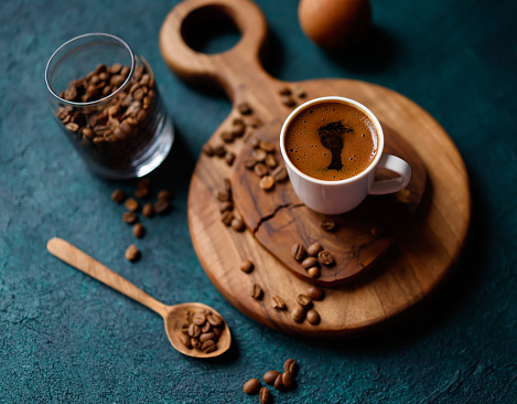 Istanbul, Turkey-April 11, 2022: A frothy Turkish coffee in a white cup on a round wooden cutting board on a dark turquoise green-blue  rough concrete floor. Coffee beans were scattered all around. Next to the cup is a jar with coffee beans and a wooden spoon. Shot with Canon EOS R5.