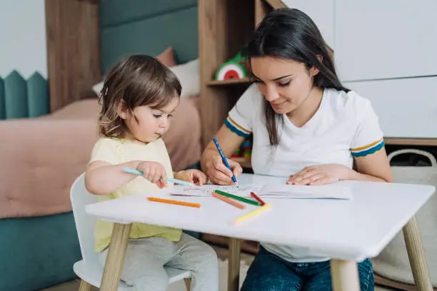 Shot of a little girl sitting at a table and coloring in a coloring book while her mother helping her. Mother teaching her daughter drawing and painting. Mom spending time with her daughter. Happy family at home.