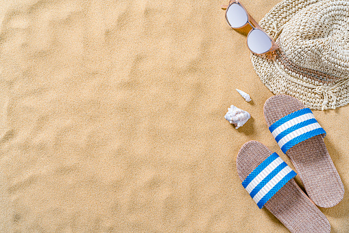 Summer backgrounds: beach sandals, sunglasses and sunhat shot from above on sand background. The composition is at the right of an horizontal frame leaving useful copy space for text and/or logo at the left. High resolution 42Mp outdoors digital capture taken with SONY A7rII and Zeiss Batis 40mm F2.0 CF lens