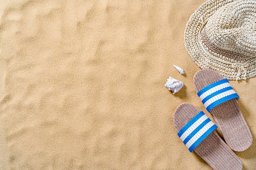 Summer backgrounds: beach sandals and sunhat shot from above on sand background. The composition is at the right of an horizontal frame leaving useful copy space for text and/or logo at the left. High resolution 42Mp outdoors digital capture taken with SONY A7rII and Zeiss Batis 40mm F2.0 CF lens
