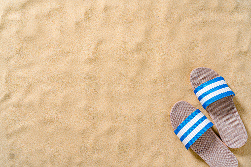 Summer backgrounds: beach sandals shot from above on sand background. The composition is at the bottom right of an horizontal frame leaving useful copy space for text and/or logo at the left. High resolution 42Mp outdoors digital capture taken with SONY A7rII and Zeiss Batis 40mm F2.0 CF lens