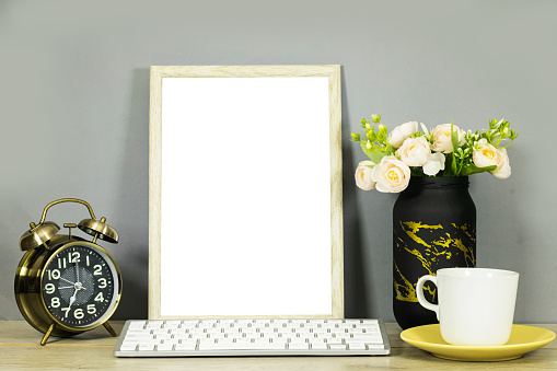 still life: empty picture frame mockup and flowers vase and cup of coffee and alarm clock, keyboard on wooden desk table. Elegant working space, home office concept