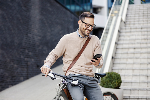 A smiling urban man sits on a bike on the street and using phone.