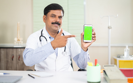 happy smiling doctor showing green screen mobile phone by pointing finger at hospital - conapt of online consultation booking, app promotion and advertisement.