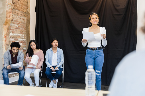The young adult woman auditions for an unrecognizable producer while other actors and actresses wait patiently for their turn.