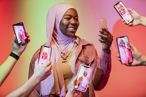 Woman wearing a hijab standing in front of a multi coloured background in the North East of England. She is holding a mobile phone and is surrounded by hands reaching towards her also holding phones with pictures of her on the screens.