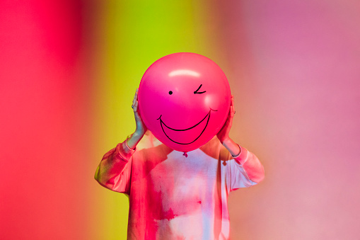 Woman standing in front of a multi coloured studio background holding a bright pink balloon with a winking face on it in front of her. She is in the North East of England.
