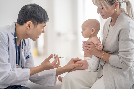A mother sits on an examination table at the doctors with her infant son propped up on her lap as he receives an immunization.  The baby is wearing only a diaper and is looking at the doctor as she injects the needle into the baby's leg.  The male doctor of Asian decent is wearing a white lab coat and is trying to distract the baby from the needle by engaging him.