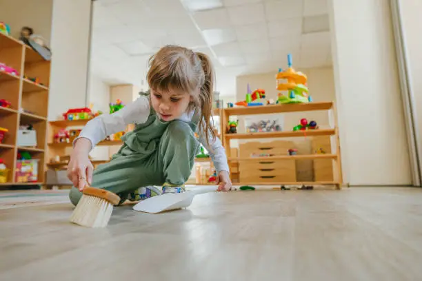 Little girl sweeping the floor using a dustpan and a broom at kindergarten or childcare centre. Montessori early development education concept.