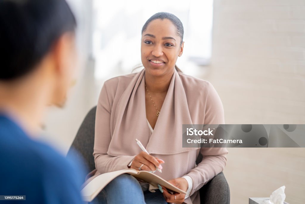 Young Man in a Therapy Session A young man of  Asian decent, sits with a therapist as he talks through his struggles.  He is dressed casually in a blue t-shirt and has his back to the camera as they talk.  The therapist is dressed professionally and has a notepad out on her lap as she takes notes throughout the session. Mental Health Professional Stock Photo