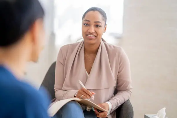 A young man of  Asian decent, sits with a therapist as he talks through his struggles.  He is dressed casually in a blue t-shirt and has his back to the camera as they talk.  The therapist is dressed professionally and has a notepad out on her lap as she takes notes throughout the session.