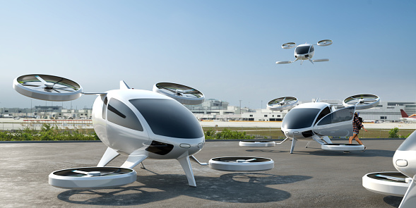 A fleet of generic white electric powered Vertical Take Off and Landing eVTOL aircraft with four rotors parked with airport buildings visible in the background. One has a door open, with a young female passenger about to get on board. Another eVTOL is in flight, coming in to land.