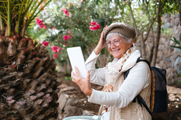 istock Cheerful senior woman with cap and backpack in outdoors excursion sitting video chatting with digital tablet.  Elderly attractive female resting in shadow of tropical woods 1395176725