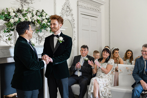 An over-the-shoulder mid-length view of two grooms who have just been pronounced husband and husband. They are holding hands and looking lovingly towards each other.