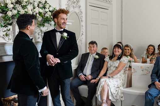 An over-the-shoulder mid-length view of two men getting married in front of their family. The man with the red hair is beaming with pride after having his wedding band placed on his hand.