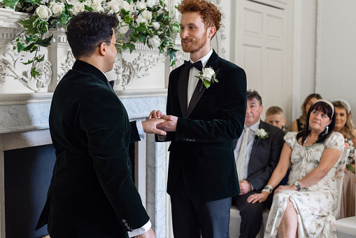 An over-the-shoulder view of two men getting married and exchanging rings with each other. The groom with the red hair is placing the ring on his partners hand,