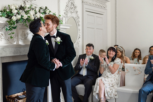 A mid-length side view of two newlyweds kissing each other after just being pronounced husband and husband. The guests can be seen clapping and cheering and celebrating the two men's big day