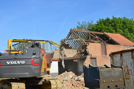 Wilsele, Vlaams-Brabant, Belgium - May 03, 2022:  Volvo excavator dismantling a car repair garage building in brick. Plastic foil hanging on the bucket