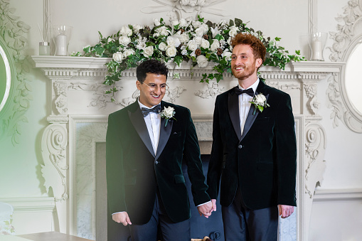 A mid-length view of a married male couple at their wedding ceremony in a public registry office. They are standing at the front in front of all of their guests. They have been pronounced husband and husband