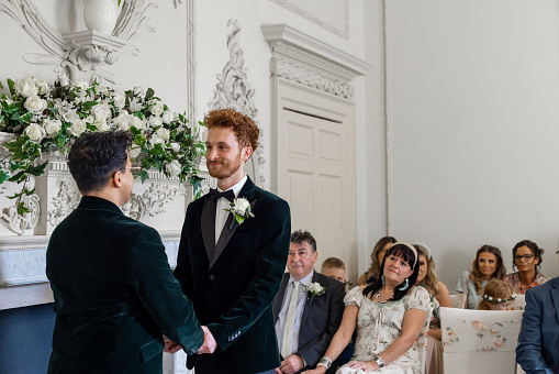 An over the shoulder view of two men getting married. They are holding hands and making their vows to one another in front of their friends and family.