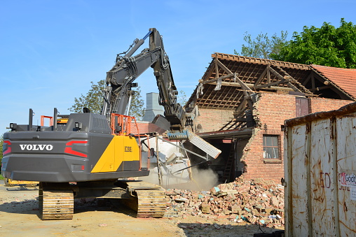 Wilsele, Vlaams-Brabant, Belgium - May 03, 2022:  Volvo excavator dismantling a car repair garage building in brick. Bulldozer grabs wall and makes dust