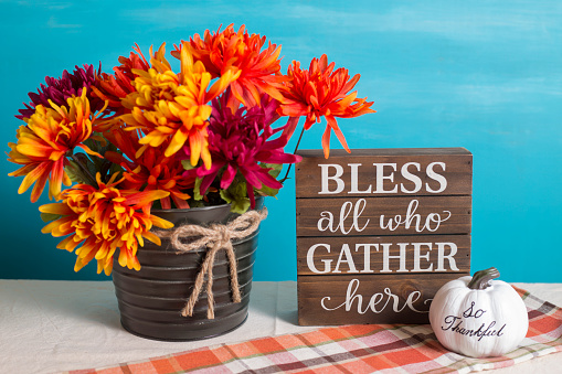 Autumn table decorations.  Bucket of orange flowers,  turquoise background and plaid table runner or cloth.  White pumpkin.  Bless all who Gather here on wooden box.