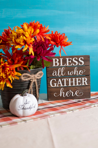 Autumn table decorations.  Bucket of orange flowers,  turquoise background and plaid table runner or cloth.  White pumpkin.  Bless all who Gather here on wooden box.