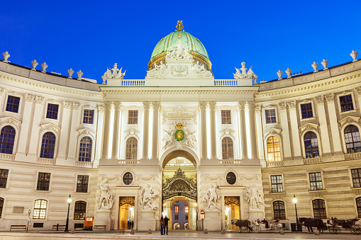 People stand on Michaelerplatz in front of St. Michaels Gate at the Hofburg Palace in Vienna, Austria at twilight.