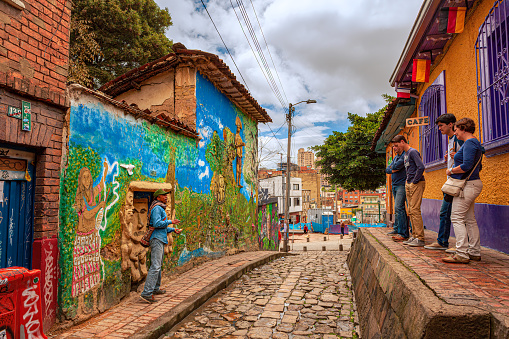 Bogota, Colombia - July 20, 2016: A tour guide explains some of the many legends of Colombia, on the cobblestoned Carrera 2, in the historic district of La Candelaria. He is carrying an Arhuaca Mochila, a bag originally made by the Arhuaca tribe of the Sierra Nevada. The capital city of Colombia, Bogota, was founded in the 16th Century in this area, by the Spanish Conquistador, Gonzalo Jiménez de Quesada. Many walls in this area are painted with either street art, or legends of the pre Colombian era, in the vibrant colours of Latin America. A small group of tourists listen to the story; behind them is a Cafe. The sky is overcast. Photo shot in the morning sunlight, on a cloudy day. Horizontal format.