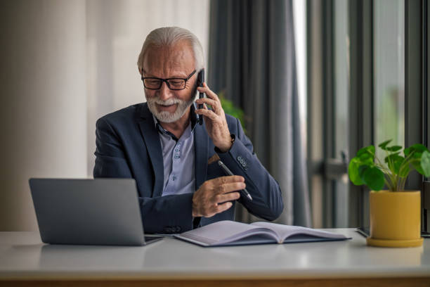 istock Smiling senior businessman talking on cellphone while working on laptop at desk 1395194675