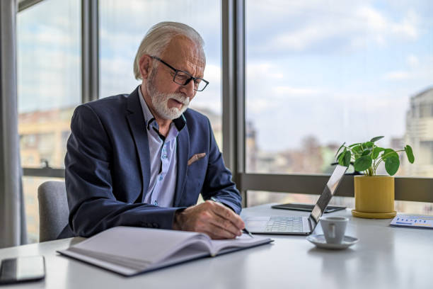istock Serious senior executive writing ideas in diary by laptop at desk in office 1395194725