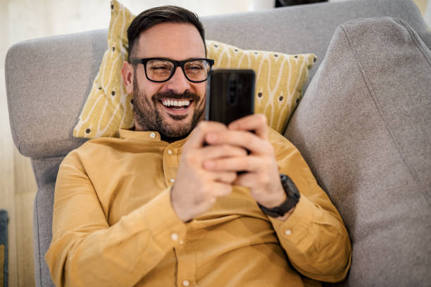 istock Cheerful young man watching video on smart phone while resting on sofa at home 1395194816