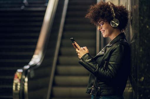 Woman listening music with wireless headphones on a smartphone during a walk in the city street. Changing songs and tracks on a mobile app. Escalator in the underground metro station in the back.