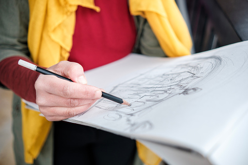 Close up of woman's hand making a drawing on a sketchbook. She is holding a pencil and we see a yellow scarf.