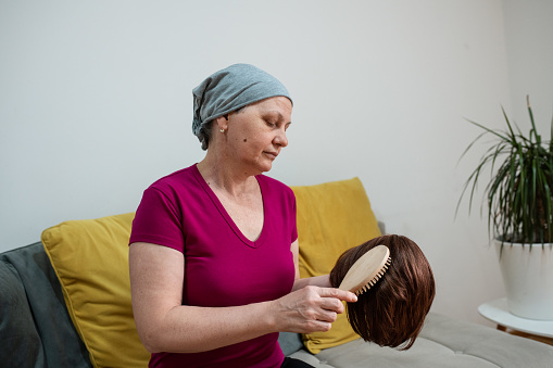 Woman suffering from breast cancer brushing her wig at home. Pancreatic cancer survivor in a headscarf. Ovarian cancer.  Pancreatic cancer. Lung cancer. Tumor. Skin cancer.  Cancer survivor.